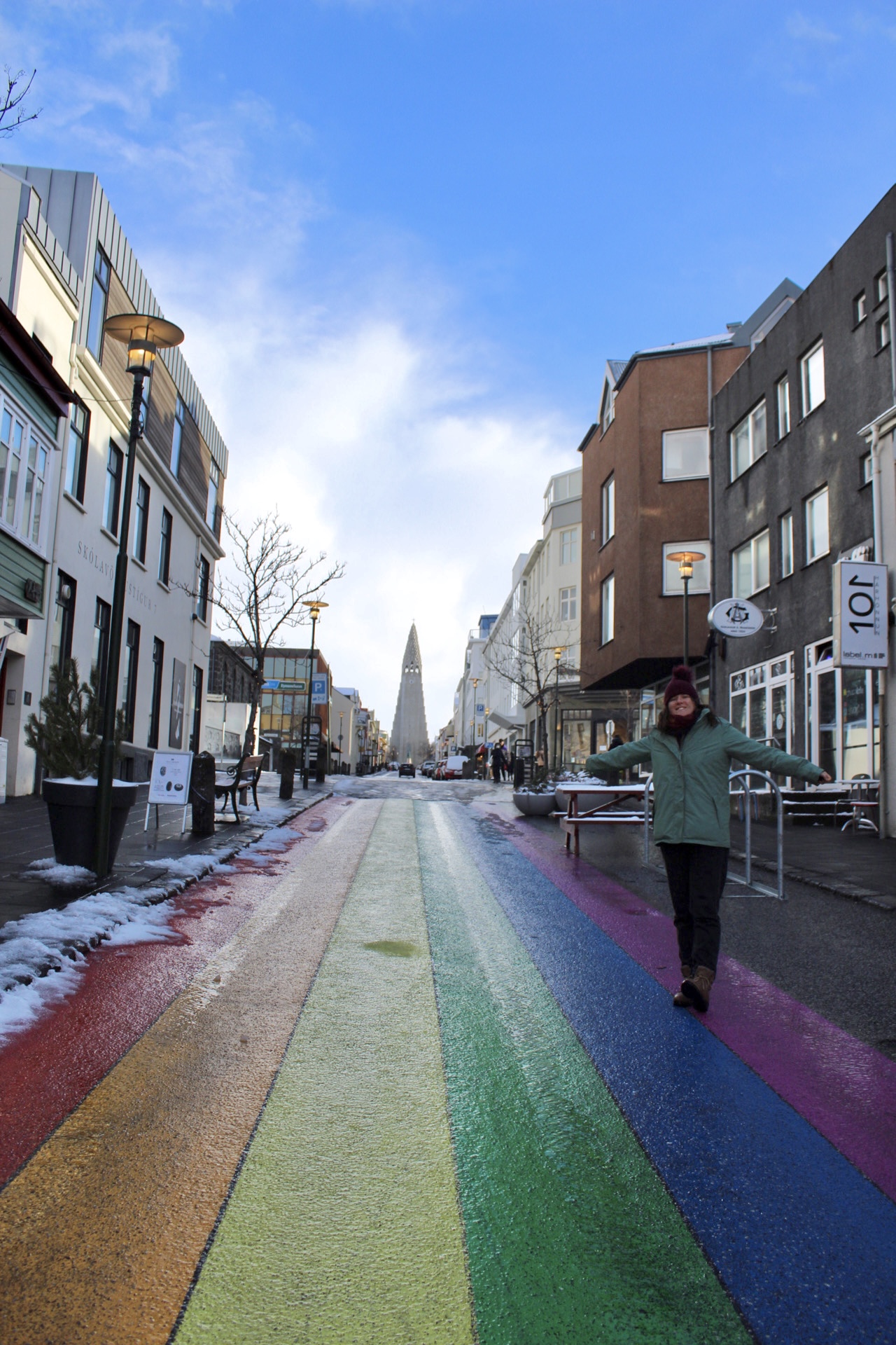a woman smiles and holds her arms out in a rainbow painted footpath looking up towards a cathedral
