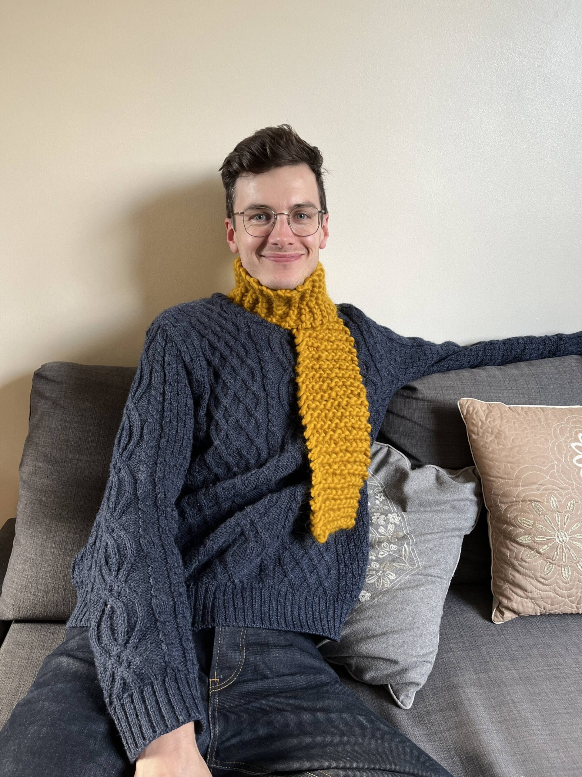 a man sits on a sofa and smiles while wearing a yellow knitted scarf