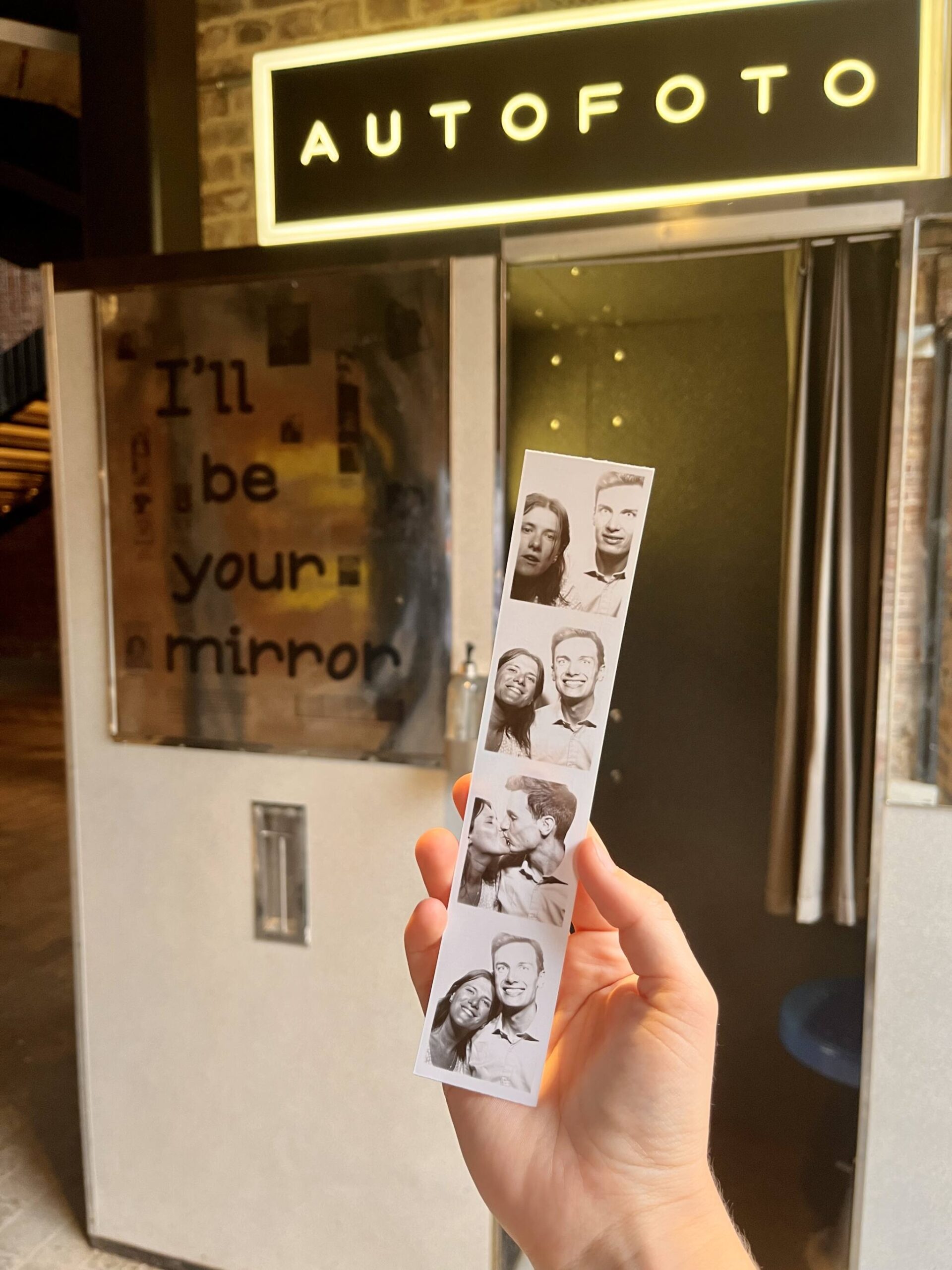 A hand holds a print out of photobooth images in front of a photobooth